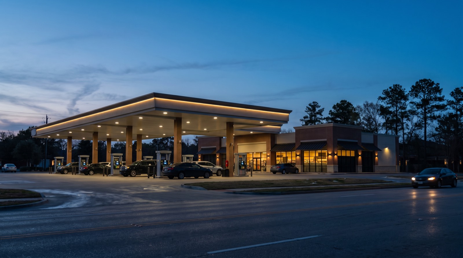 Illustrative image, not the actual property. A fuel station canopy and small strip center with lit storefronts in a Texas suburb at dusk
