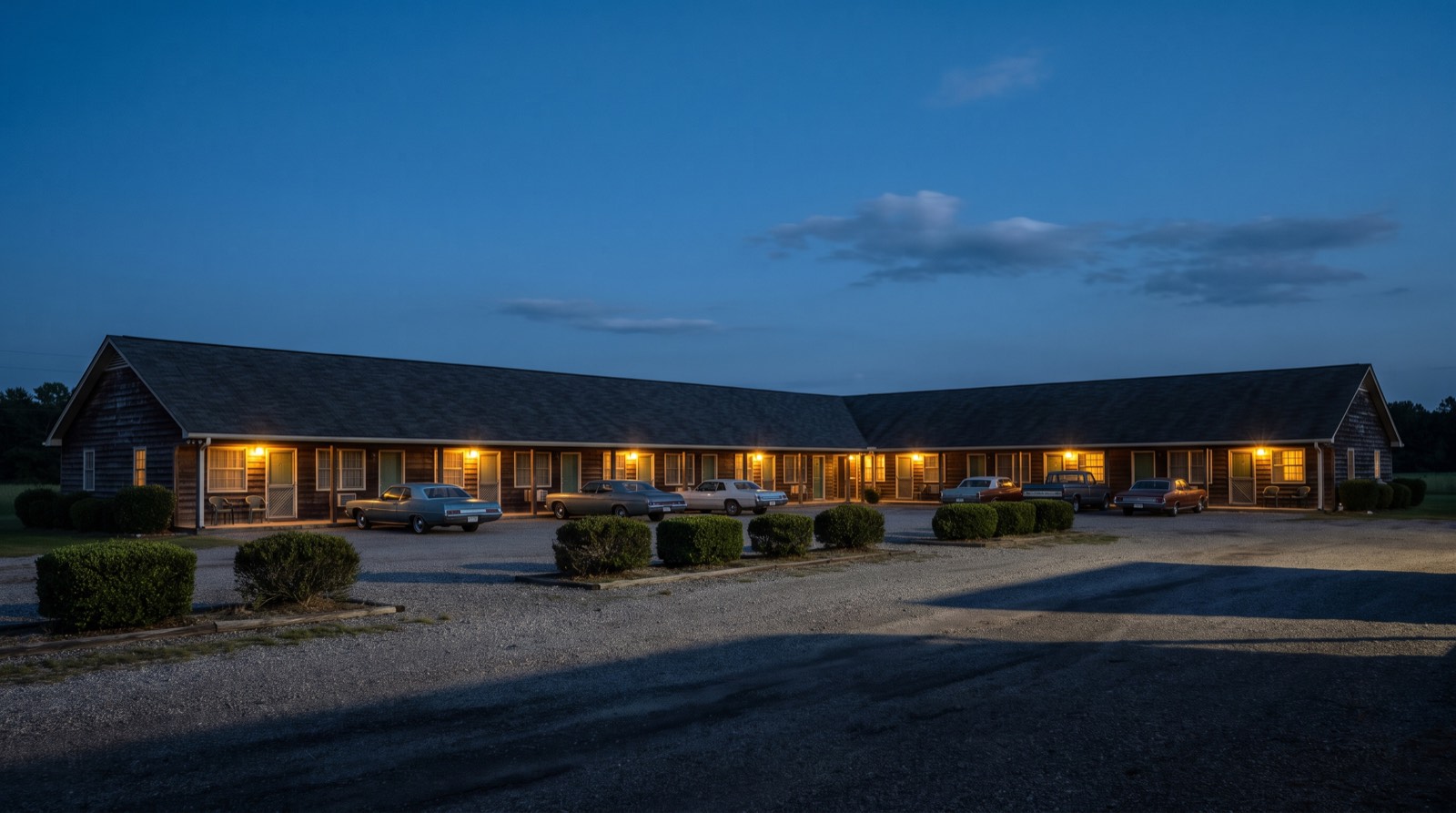 Illustrative image, not the actual property. A long single-story roadside motel with lit room windows and cars in the lot at dusk
